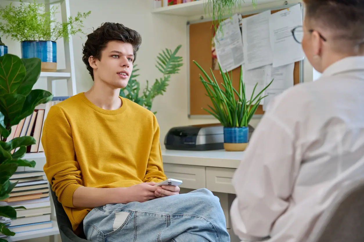 Two men converse, one sitting in a chair while the other stands beside him